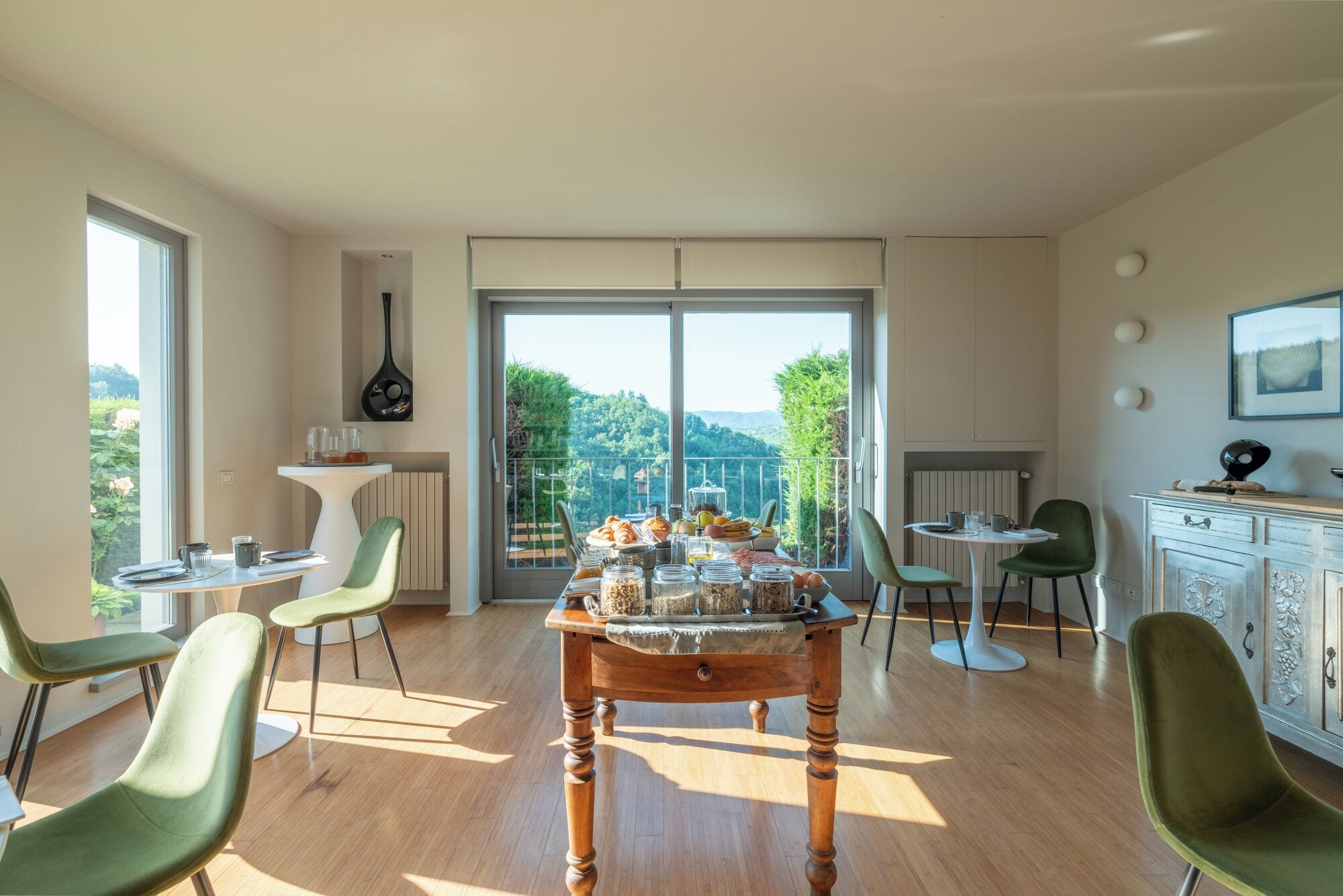 A spacious dining area with a large table set for breakfast, surrounded by modern chairs, and a view of lush greenery through large windows.