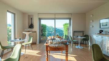 A spacious dining area with a large table set for breakfast, surrounded by modern chairs, and a view of lush greenery through large windows.