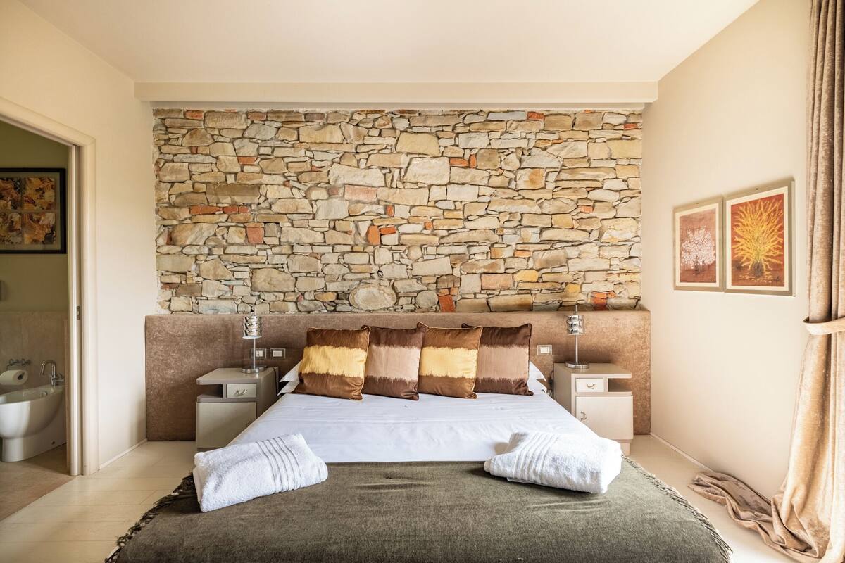 A bedroom with a stone wall, a bed with multiple pillows, two bedside tables, and a nightstand.