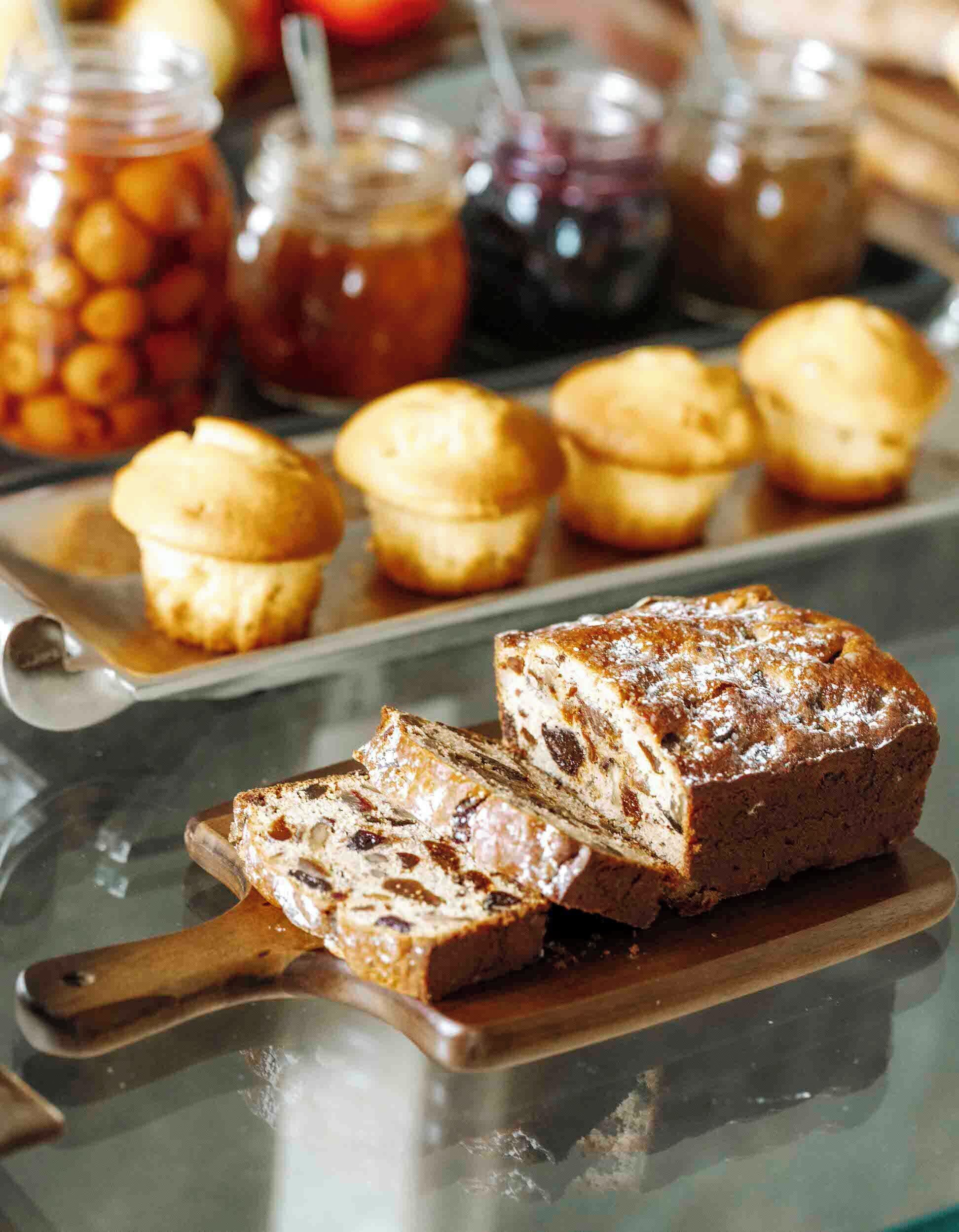 Une miche de gâteau aux fruits et aux raisins secs, posée sur une planche à découper en bois, accompagnée d’un plateau de muffins et de pots de confitures.