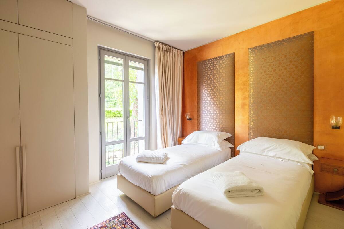 A hotel room with two beds, a large window with curtains, a wardrobe, and a patterned wall.
