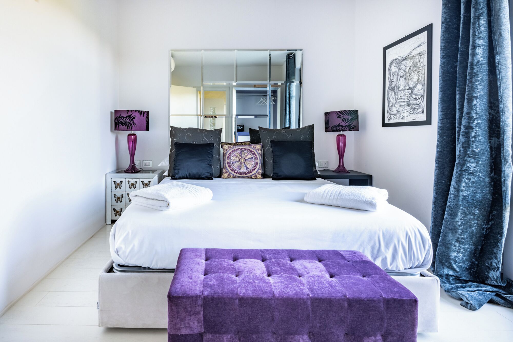 A modern bedroom with a large bed, two bedside tables, a mirror, and a purple ottoman.