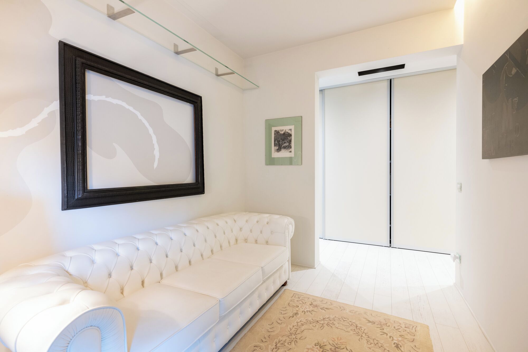 A white sofa in a room with a glass shelf, a framed picture, and a door.
