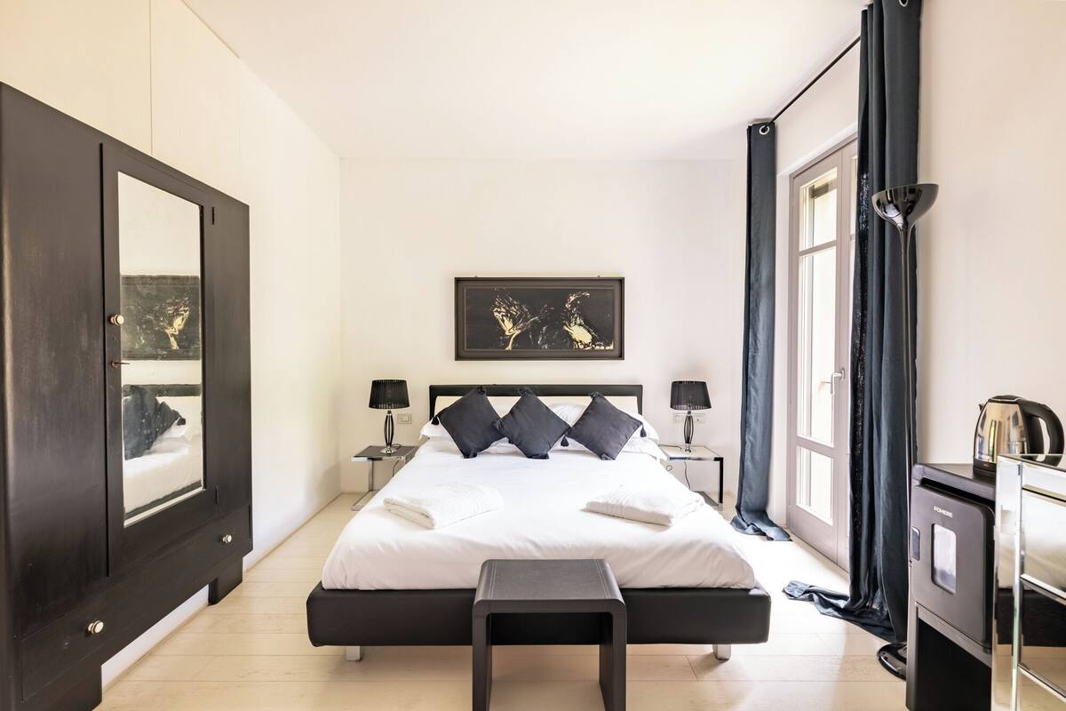 A modern bedroom with a large bed, two bedside tables with lamps, a black bench, a framed artwork above the bed, and a wardrobe with a mirror.
