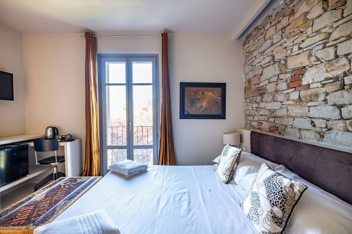 A hotel room with a bed, a desk with a computer, a stone wall, and a window with curtains.