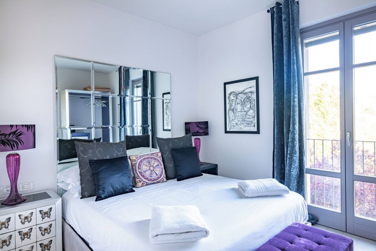 A bedroom with a large bed, a dresser, a mirror, and a window with curtains.