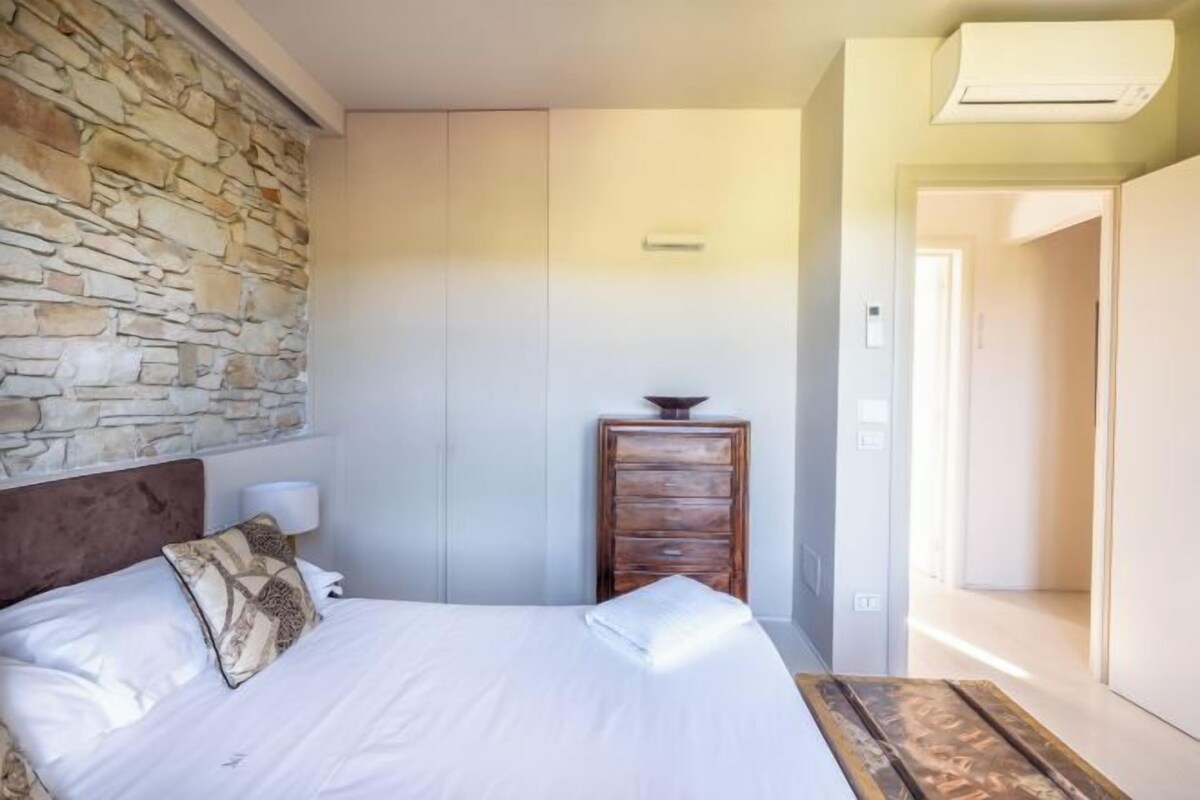 Une chambre avec un mur en pierre, un lit recouvert de draps blancs, une commode en bois et une table de chevet avec une lampe.