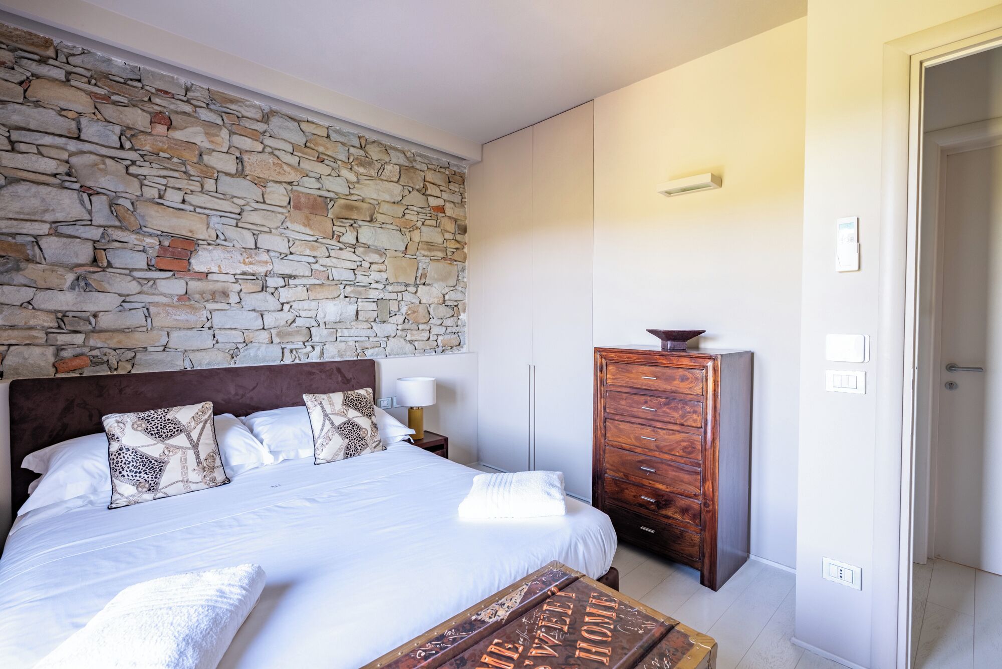 A bedroom with a stone wall, a wooden dresser, a bed with patterned pillows, and a bedside table with a lamp.