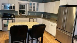 A kitchen with white cabinets, a stainless steel refrigerator, a microwave, a stove, and a granite countertop. - Charming Falmouth Heights Steps to Beach (Falmouth)