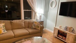 A living room with a beige sofa, a glass coffee table, a TV, and a bookshelf. - Charming Falmouth Heights Steps to Beach (Falmouth)