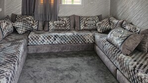 Ein modernes Wohnzimmer mit einem Ecksofa, einem großen Fenster mit Jalousien und einer Deckenleuchte.
