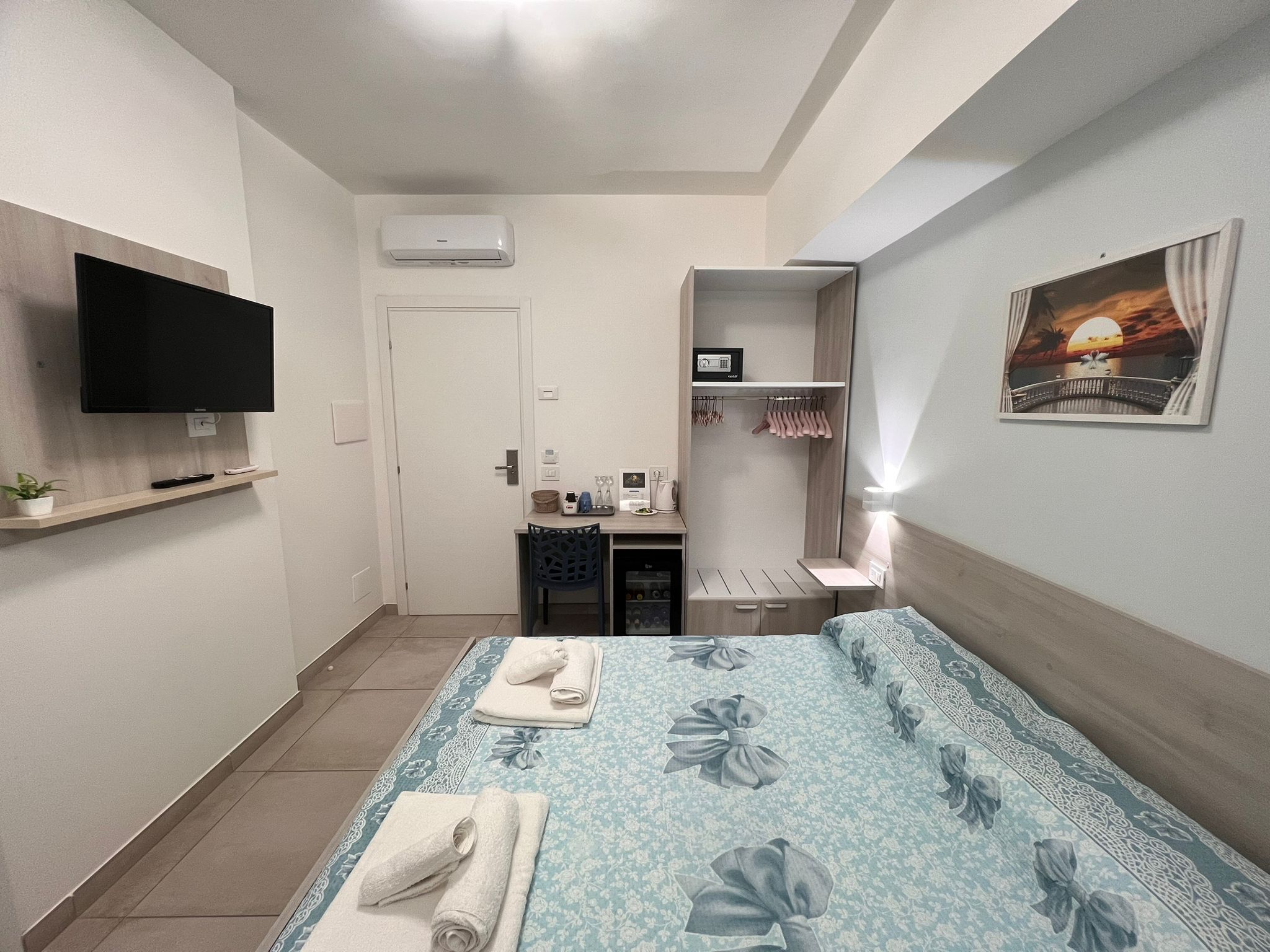 A hotel room with a bed, a TV, a desk, a wardrobe, and a painting on the wall.