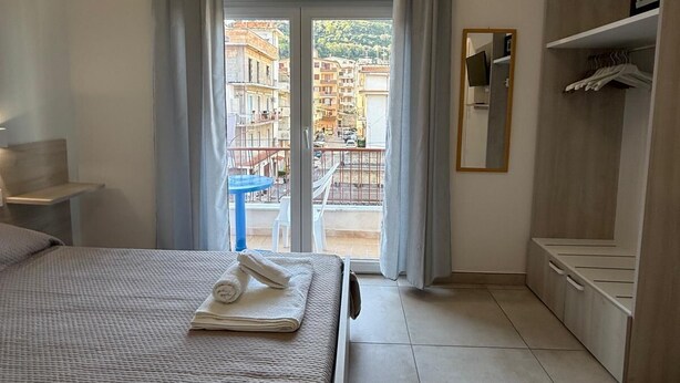 Habitación de hotel con cama, armario, espejo y balcón con vistas a edificios.