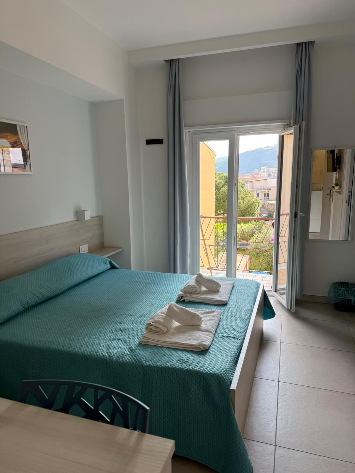 A hotel room with a bed, a table, a chair, a balcony with a view, and a door leading to the outside.