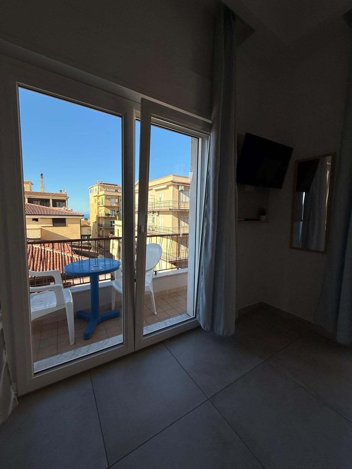 Una habitación con puerta de cristal corredera que da a un balcón, una pequeña mesa redonda con sillas, un televisor montado en la pared y suelo de baldosas.