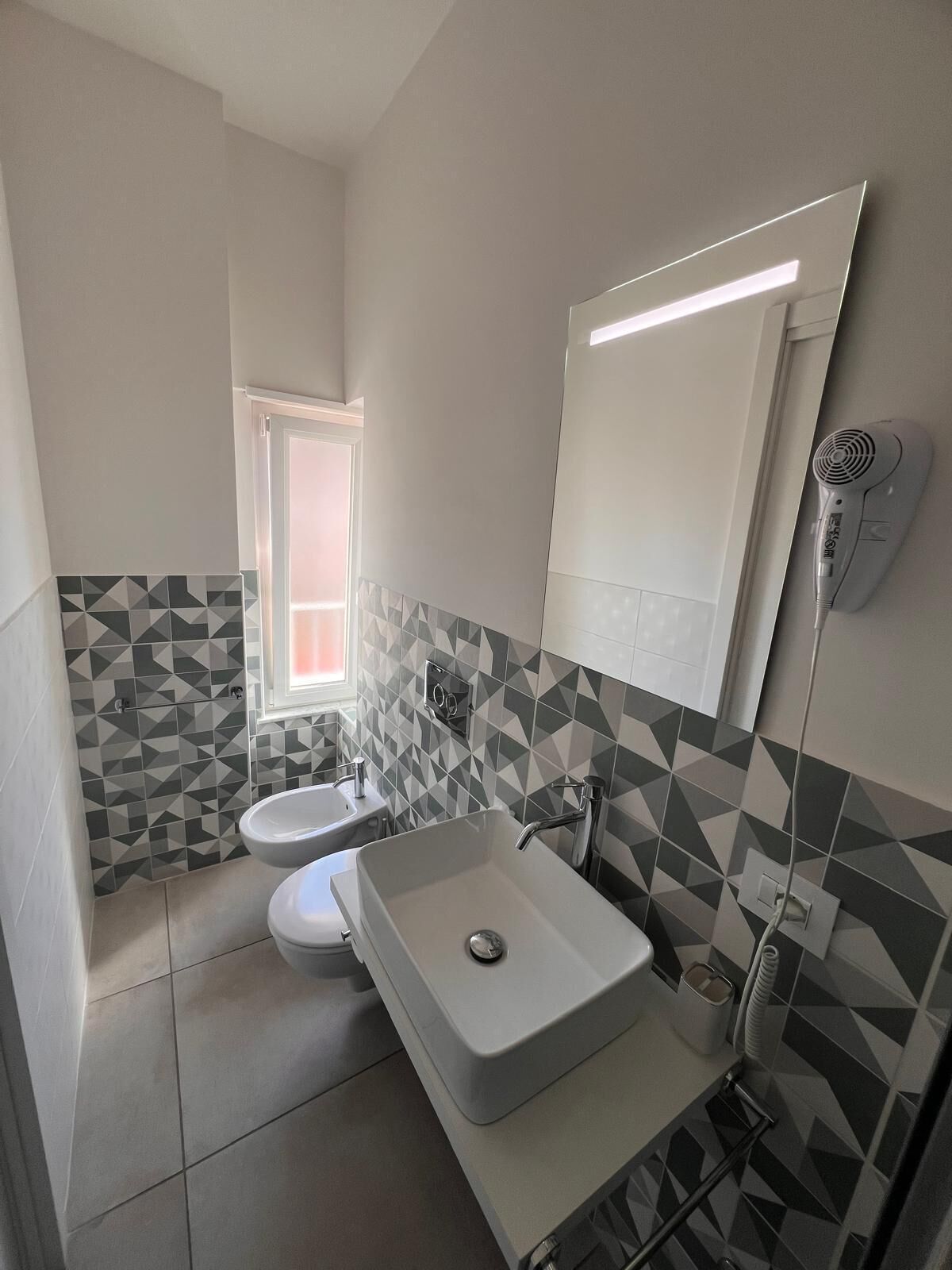 Un baño moderno con un salpicadero de azulejos geométricos, un espejo grande, un secador de pelo montado en la pared y una ventana que proporciona luz natural.