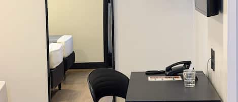 A compact hotel room with a mirrored wardrobe, a desk with a chair, a hanging coat rack, a television, and a trash bin.