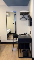 Quarto de hotel compacto com guarda-roupa espelhado, escrivaninha com cadeira, cabideiro, televisão e lixeira.