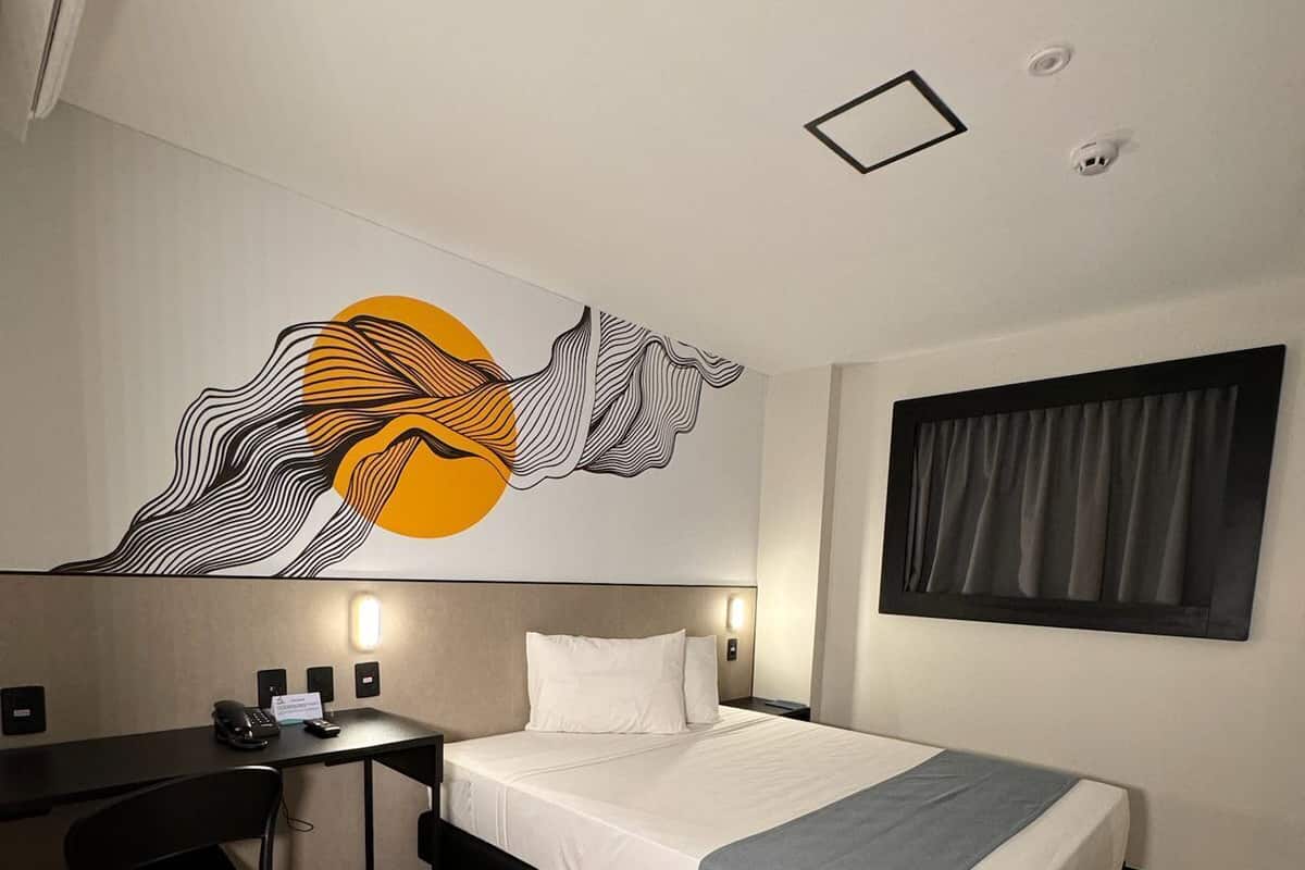 A hotel room with a bed, desk, chair, and a wall-mounted artwork.