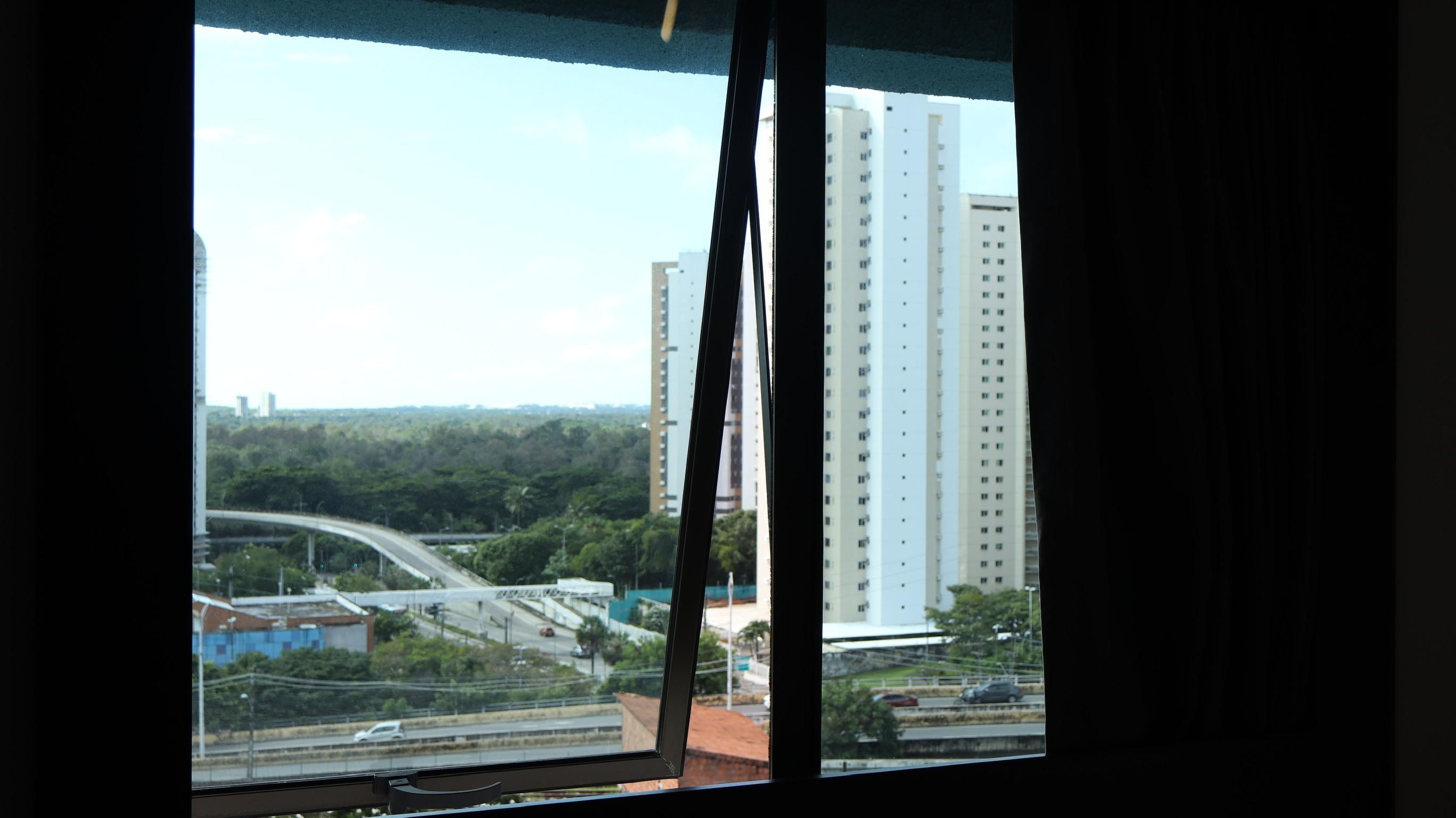 Vista pela janela, com um edifício alto ao fundo, uma ponte e vegetação.