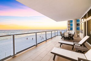 Un balcón con vistas a la playa y al océano al atardecer. Hay sillones de descanso y una mesa en el balcón.