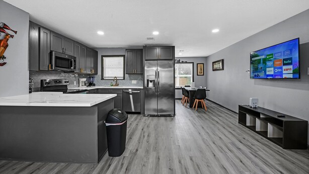 A modern kitchen with grey cabinets, a stainless steel refrigerator, and a flat-screen TV mounted on the wall.