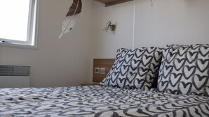 Une chambre à coucher avec une couvre-lit à motifs, une table de chevet, une lampe fixée au mur et une fenêtre avec des stores.