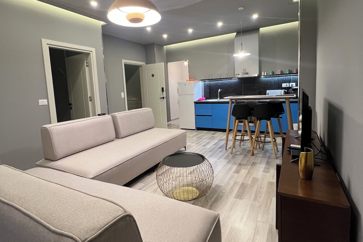Een moderne woonkamer met een bank, een salontafel, een televisie en een keuken op de achtergrond.