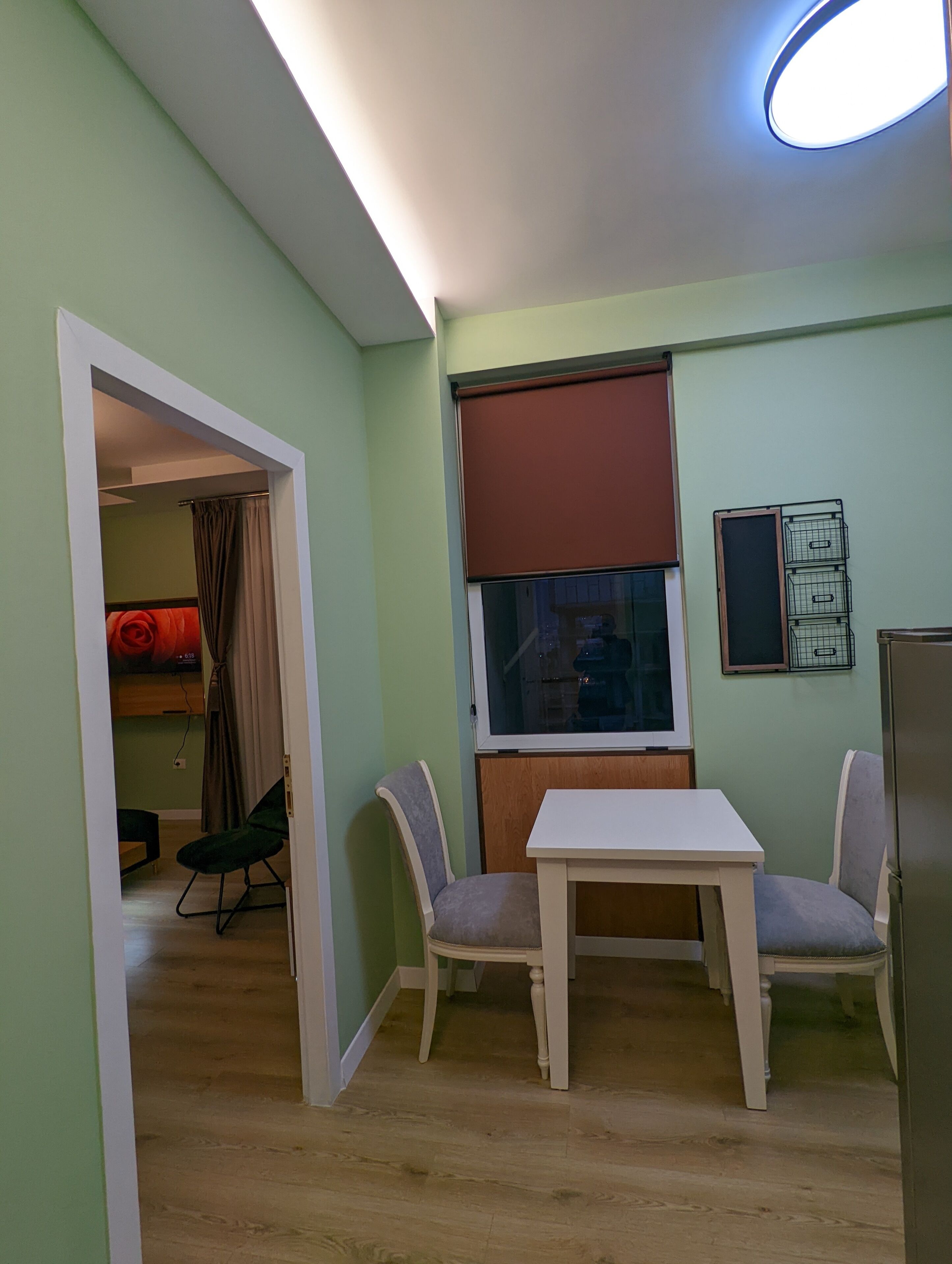 Una habitación pequeña y bien iluminada con una mesa de comedor y dos sillas, una ventana con persiana, y una puerta que da a otra habitación.