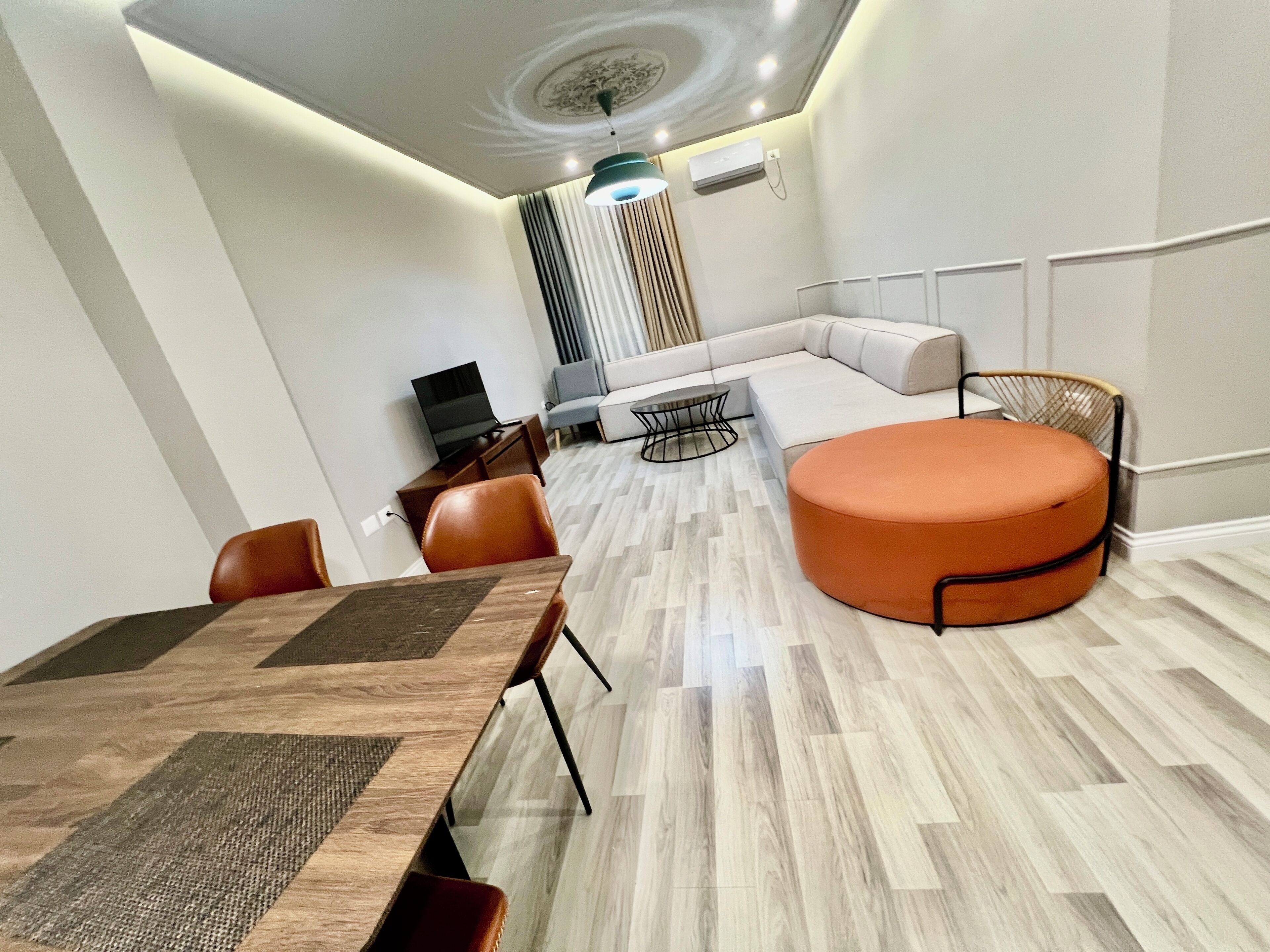 A modern living room with a sectional sofa, a round orange ottoman, a dining table with chairs, and a TV.