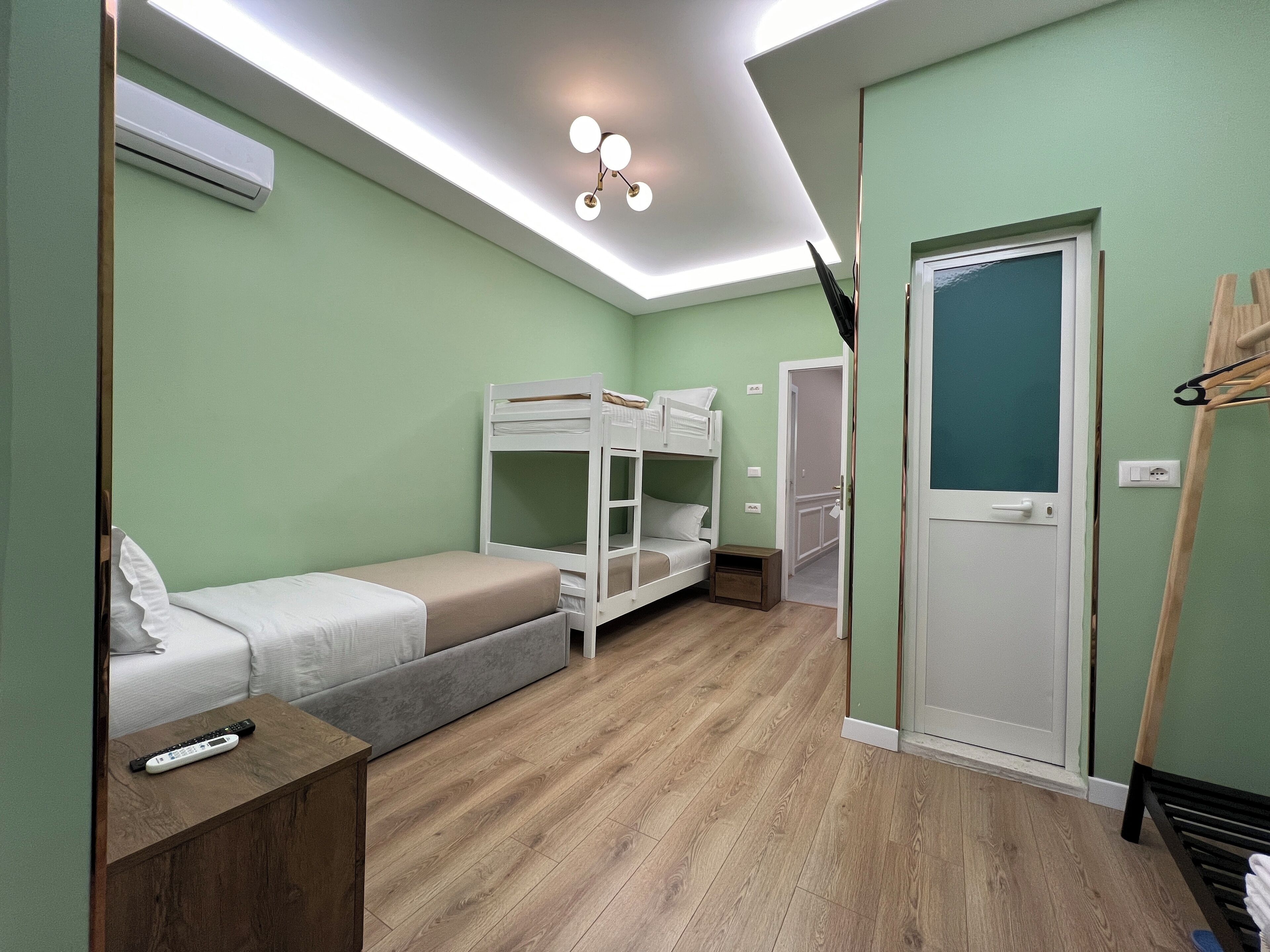 A room with bunk beds, a bedside table, a small bedside table, a door, and a light fixture.