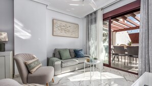 Ein modernes Wohnzimmer mit einer Couch, einem Sessel und einem kleinen runden Tisch. Ein großes Fenster mit Vorhängen bietet Blick auf eine Terrasse.