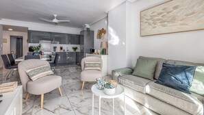 Ein modernes Wohnzimmer mit einem grauen Sofa, zwei Sesseln, einem Couchtisch und einem Essbereich mit einer Küche im Hintergrund.