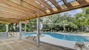 En træterrasse med swimmingpool, liggestole og et pergolaoverdækket område.
