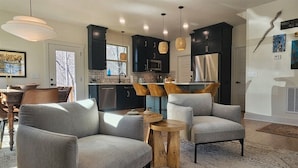 A modern living room with grey armchairs, a wooden coffee table, and a kitchen in the background.