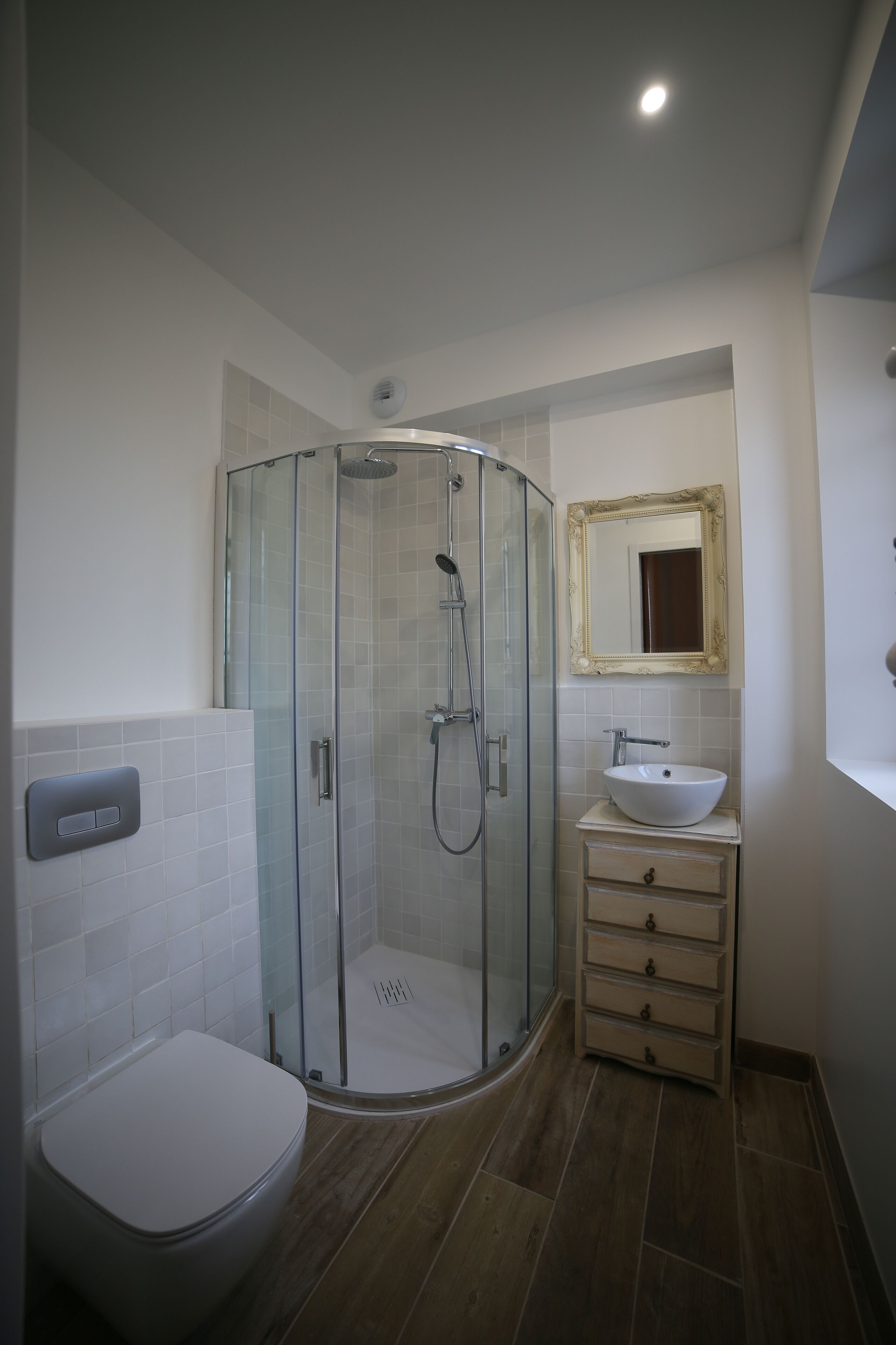 A bathroom with a glass-enclosed shower, a white toilet, a sink with a mirror, and a wooden floor.