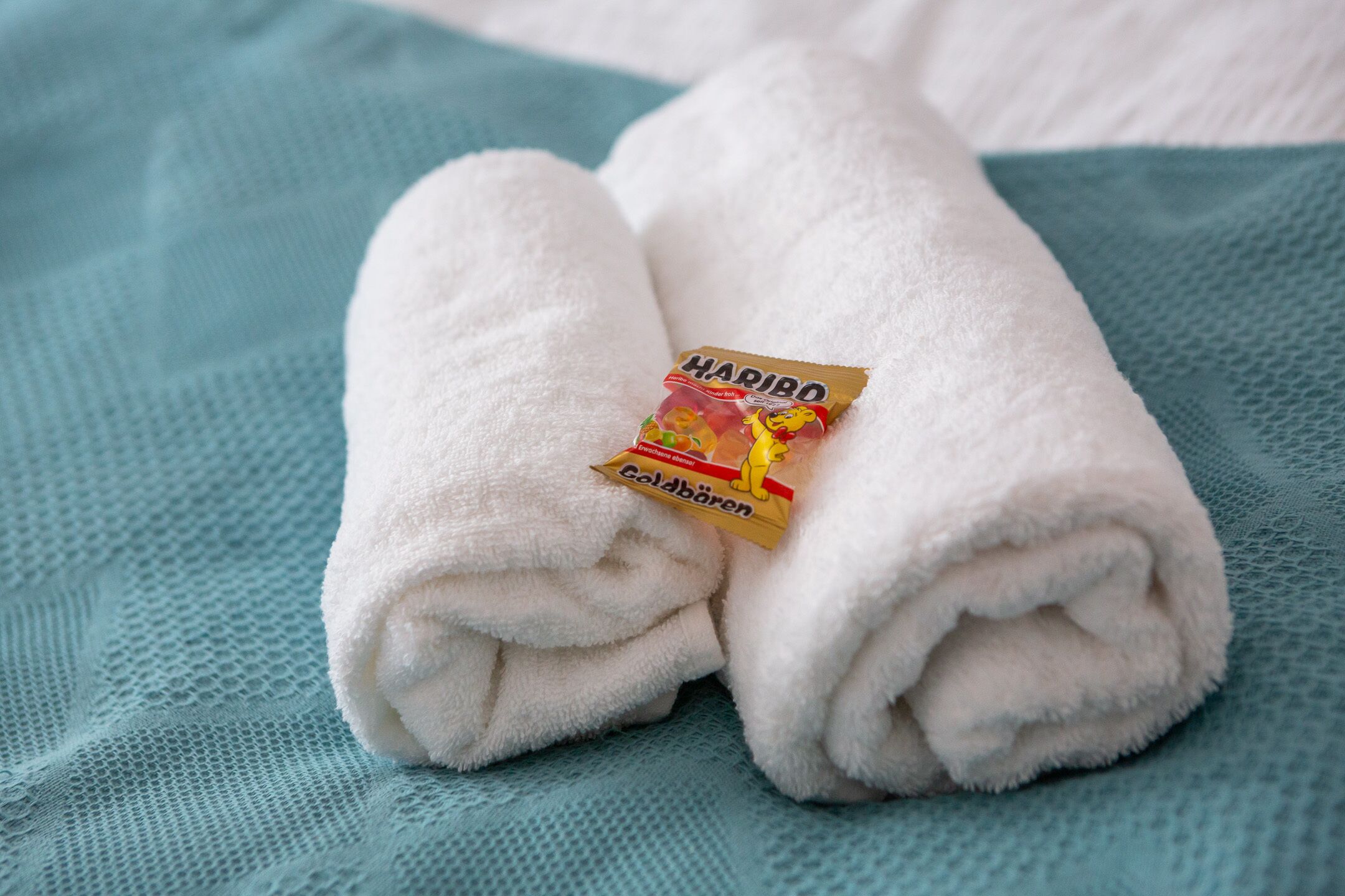 En prydligt vikt, vit handduk tillsammans med ett paket Haribo Coldbären på en strukturerad, turkosfärgad yta.