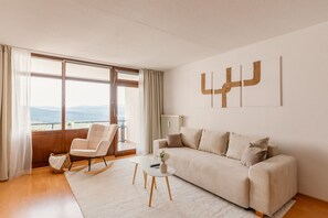 Ein modernes Wohnzimmer mit einer Couch, einem Schaukelstuhl, einem Couchtisch und Blick ins Freie durch große Fenster.