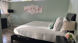 A bedroom with a bed, a nightstand with a lamp, a wall art piece, and a window with curtains. - Walk across the street to Disneyland parking. Fireworks show from front porch! (Anaheim)
