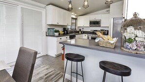 A modern kitchen with white cabinets, a central island with stools, pendant lighting, and a decorative birdcage. - Walk across the street to Disneyland parking. Fireworks show from front porch! (Anaheim)