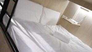 Un lit superposé avec une literie blanche et une tête de lit en bois. Il y a une petite étagère avec un miroir et une lampe fixée au mur au-dessus du lit.