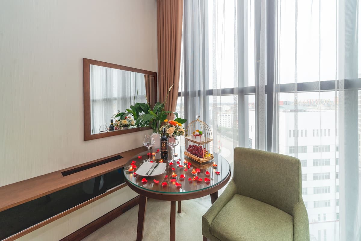 Habitación de hotel con una mesa de vidrio redonda dispuesta para comer, un sillón verde, un ventanal con vista a la ciudad y un espejo en la pared.