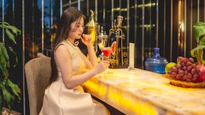 A woman in a white dress sitting at a table with a martini glass, surrounded by decorative bottles and a fruit bowl. - Bien Bac Hotel (Mong Cai)