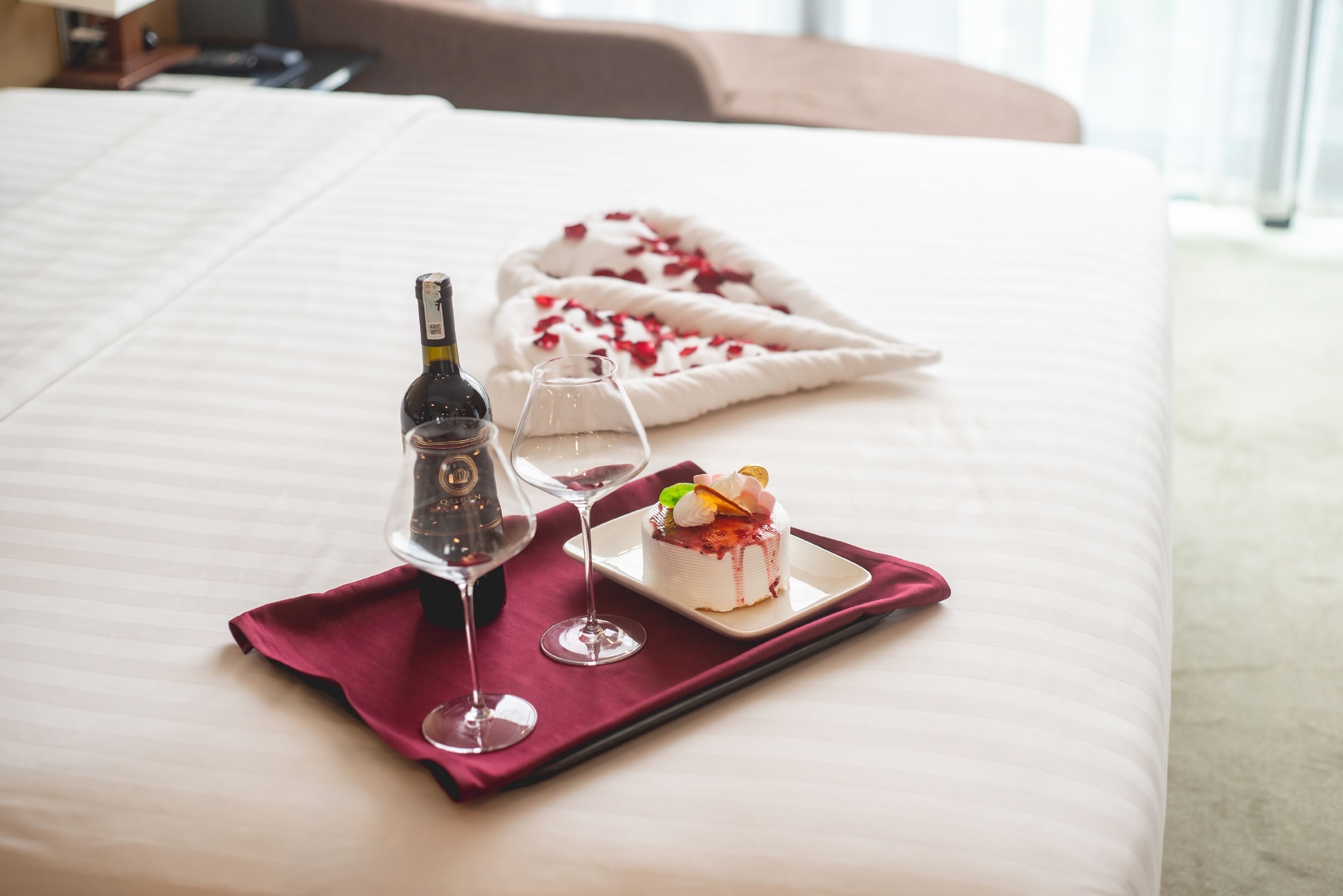 A neatly made bed with a tray holding a bottle of champagne, a glass, and a slice of cake on a red cloth.