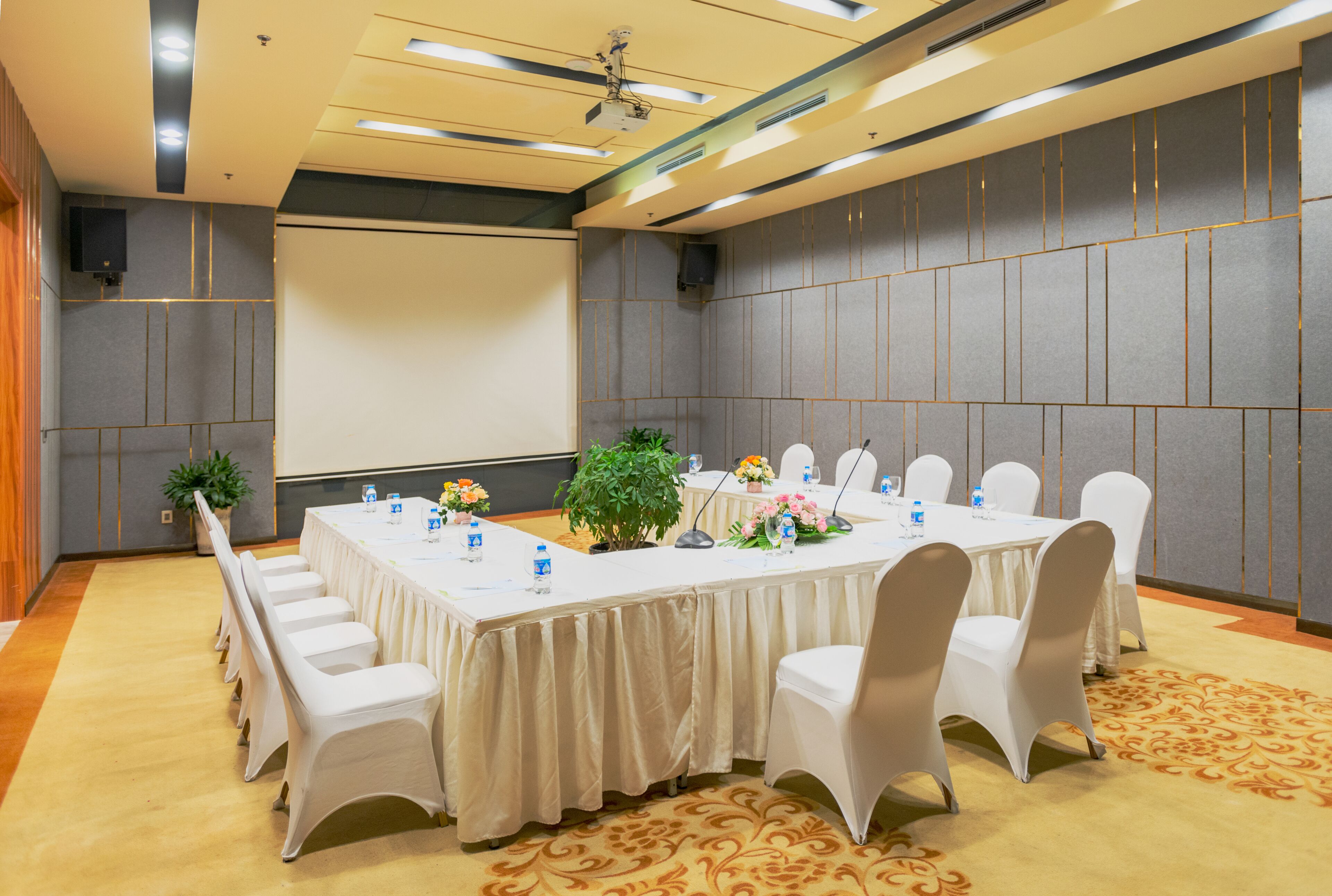 Sala de conferência com uma mesa comprida, cadeiras brancas e tela de projetor.