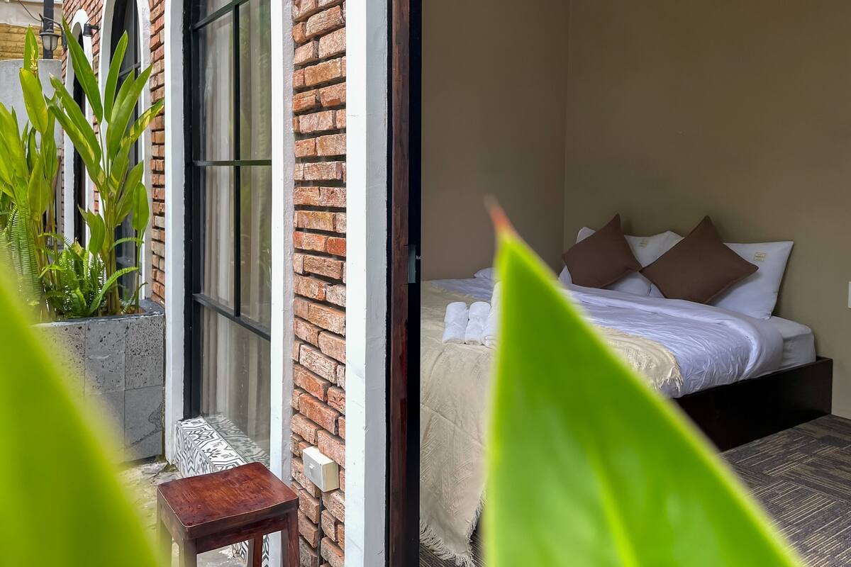 A room with a bed, a small table, and a window with a view of greenery.