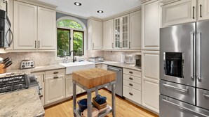 A modern kitchen with white cabinets, stainless steel appliances, and a wooden cutting board on a metal stand. - Mystic on Broadway! Downtown. Your Ultimate Mystic Getaway. Just Steps to it All (Stonington)