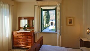 A bedroom with a wooden dresser, a bed, a window with a view of a building and greenery, and a framed picture on the wall. - Il Castello (Barbarano Vicentino)