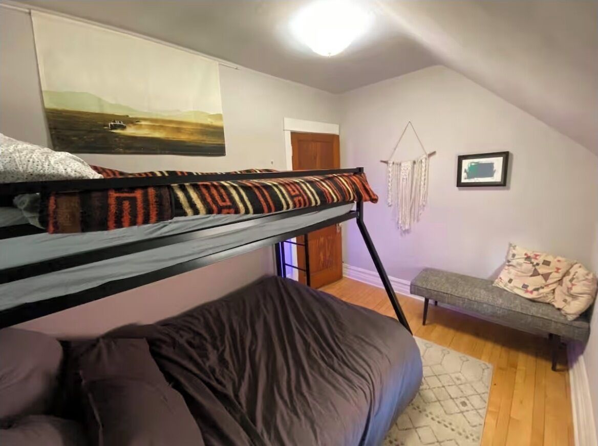 A bunk bed with a patterned blanket, a small bench, a macrame wall hanging, and a framed picture.