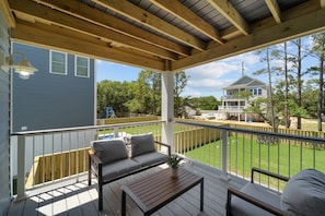 A covered deck with a sofa, a table, and a railing overlooking a backyard.